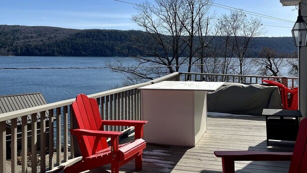 A wooden deck with red Adirondack chairs, a covered seating area, and a view of a lake and forested hills. - Lion View ~ Otsego Lake ~ Sleeps up to 10 (Cooperstown)