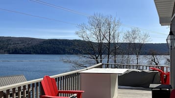 Une terrasse en bois avec des fauteuils Adirondack rouges, un espace salon couvert et une vue sur un lac et des collines boisées.