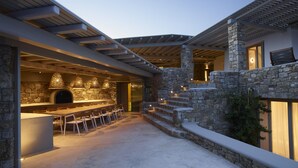 Una cocina moderna al aire libre con una pared de piedra, una mesa larga para comer y luces colgantes.