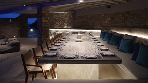 Una larga mesa de comedor dispuesta para una comida, con sillas a ambos lados, en un moderno espacio exterior con paredes de piedra y un banco de piedra.