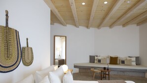 Un dormitorio moderno con techo de madera, una cama grande con ropa blanca, una mesita de noche con un libro y un taburete pequeño de madera.