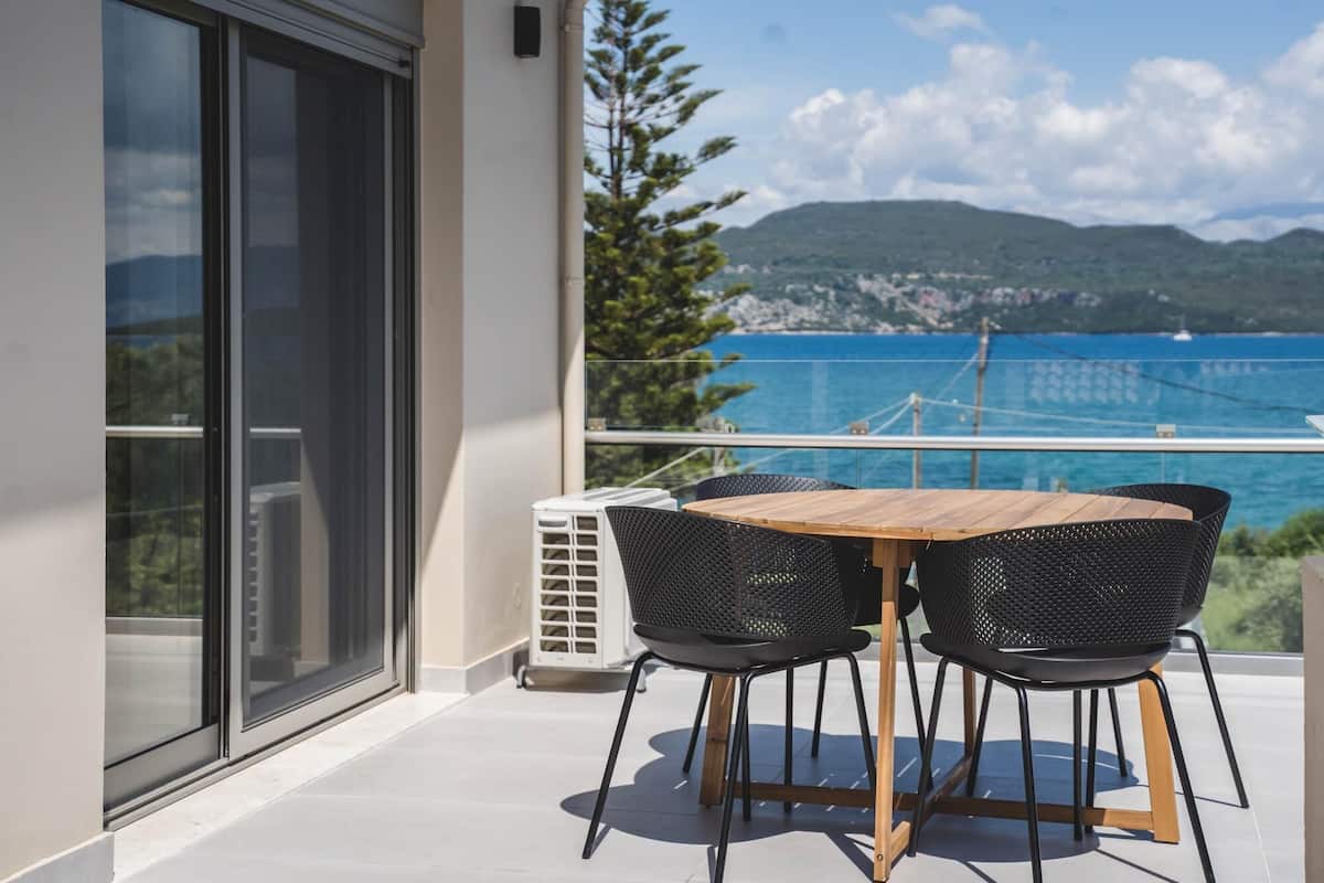 A balcony with a wooden table and black chairs, overlooking a coastal view with mountains and a bridge.