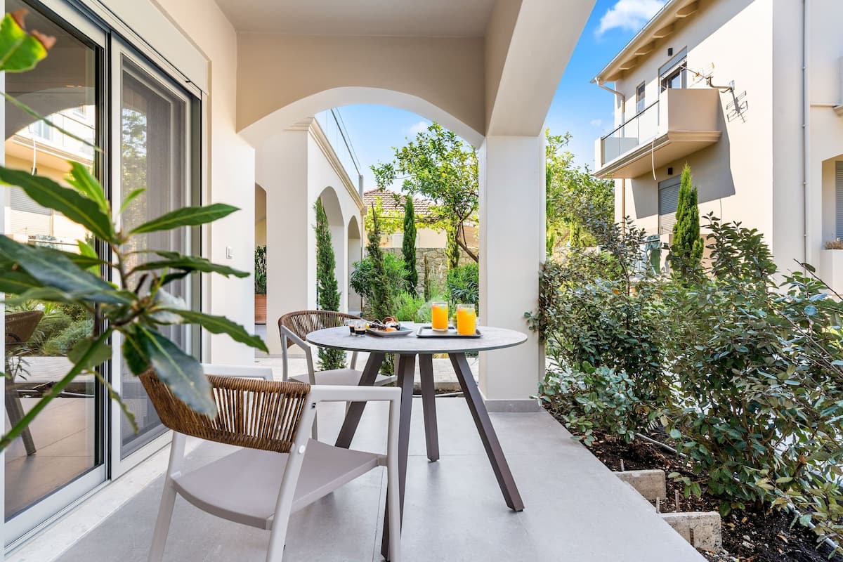 A patio with a round table and chairs, a glass door, and a view of a building and greenery.