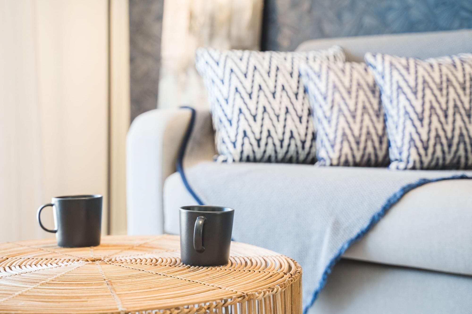 A room with a grey sofa, two patterned pillows, and two black mugs on a woven round table.