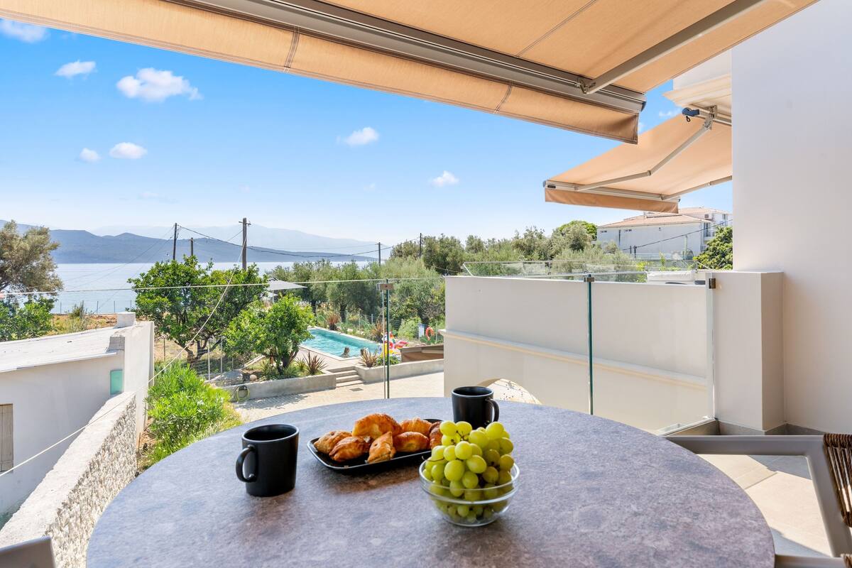 A balcony with a table set with croissants, grapes, and two black mugs, overlooking a pool and mountains.