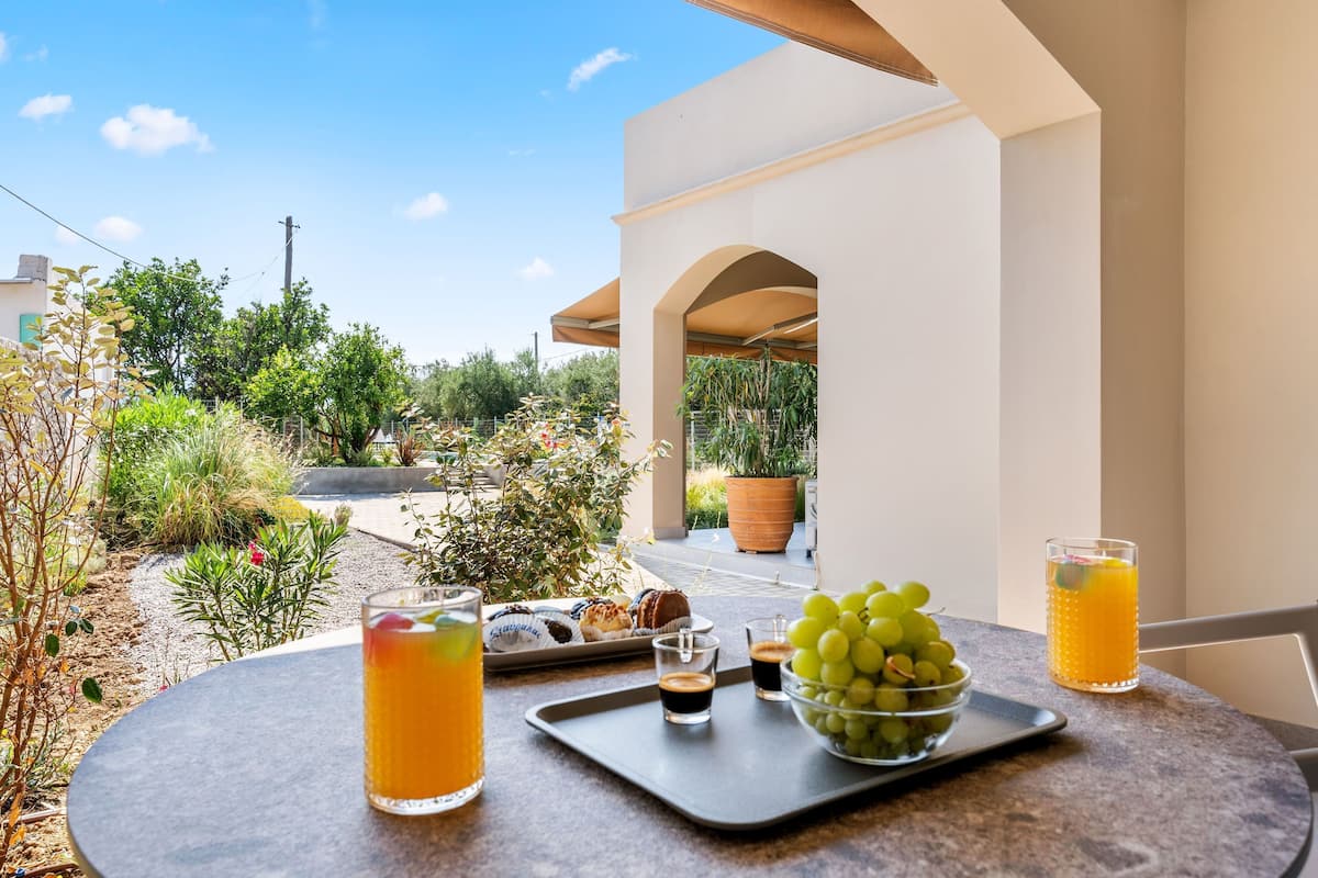 A table with a tray of pastries, a bowl of grapes, and glasses of orange juice on a balcony overlooking a garden.