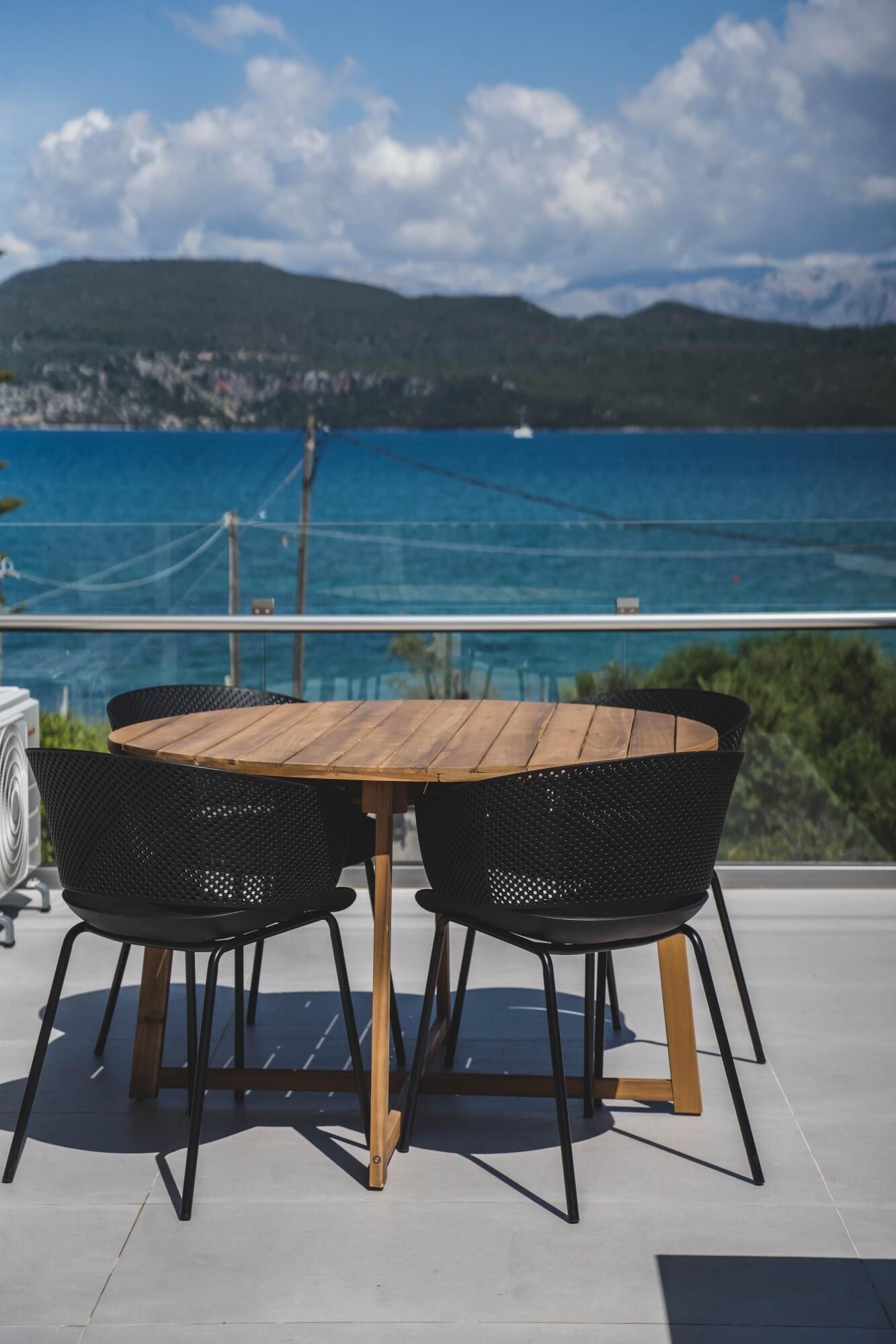 A round wooden table with four black chairs, set on a balcony overlooking a body of water and hills.