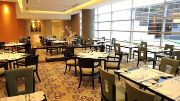 A hotel dining area with multiple tables set for dining, large windows providing natural light, and a patterned carpet.