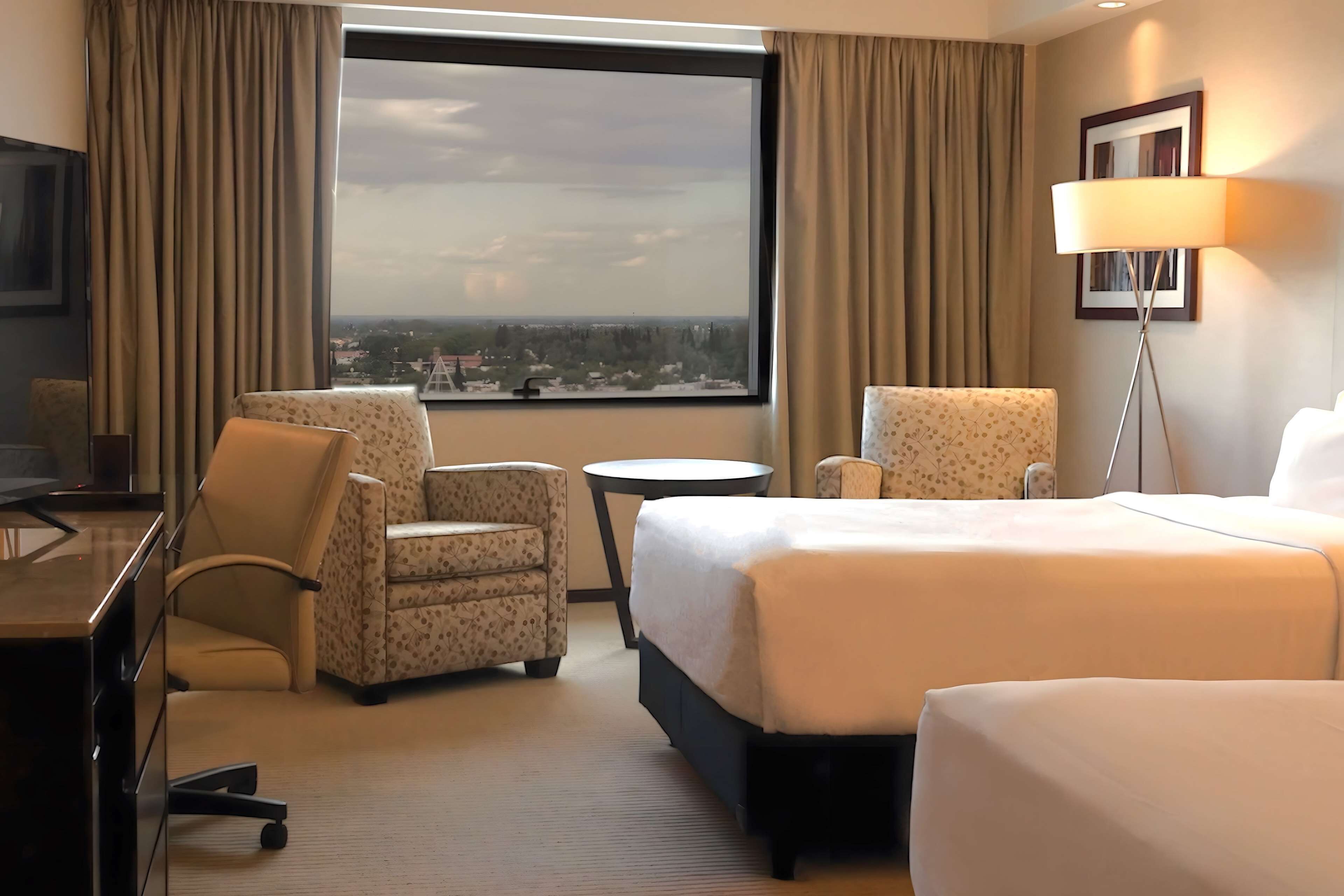 A hotel room with a large bed, two armchairs, a desk, a small table, and a view of the city through a large window.