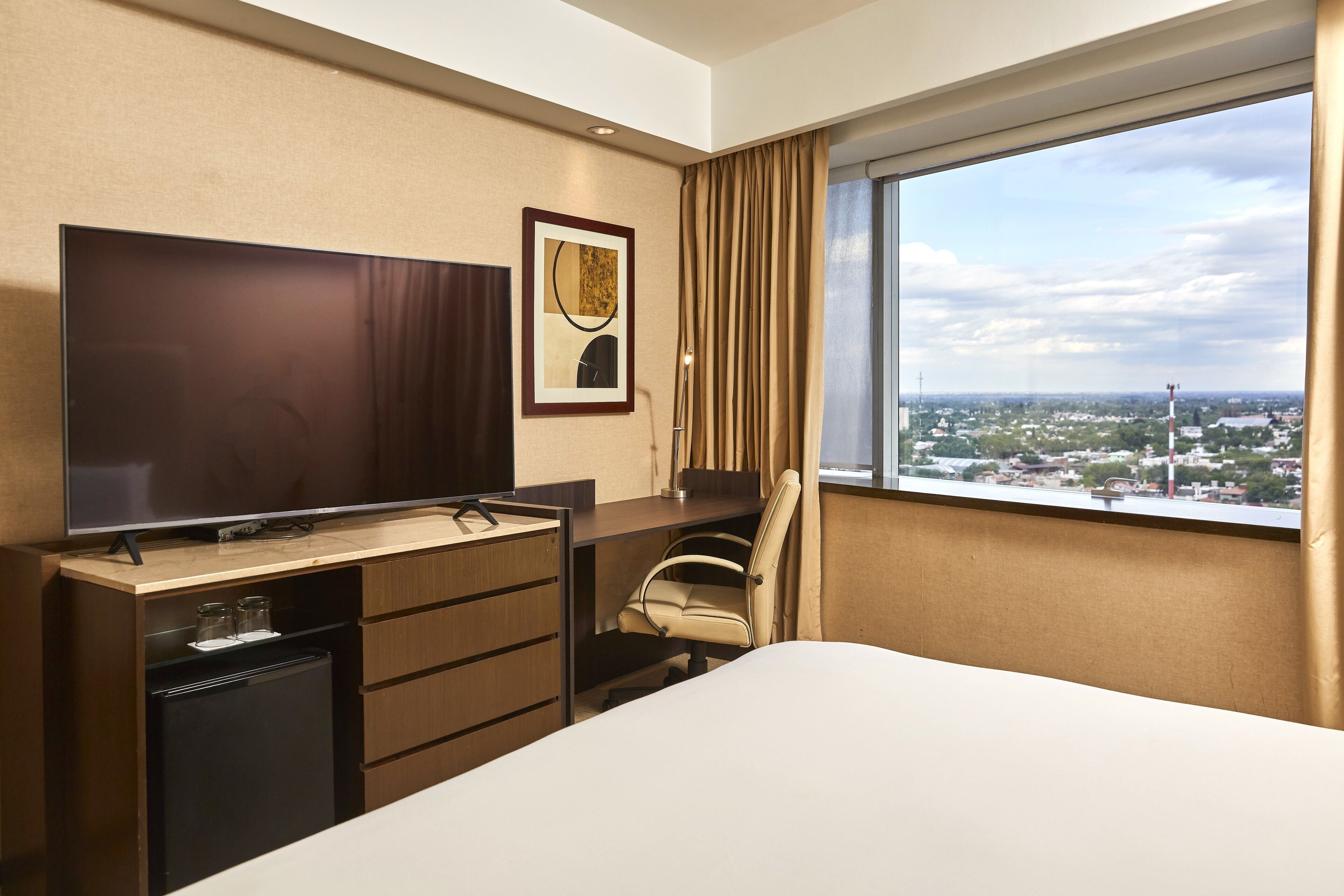 A hotel room with a bed, a desk with a television, a chair, a small refrigerator, and a window with a city view.