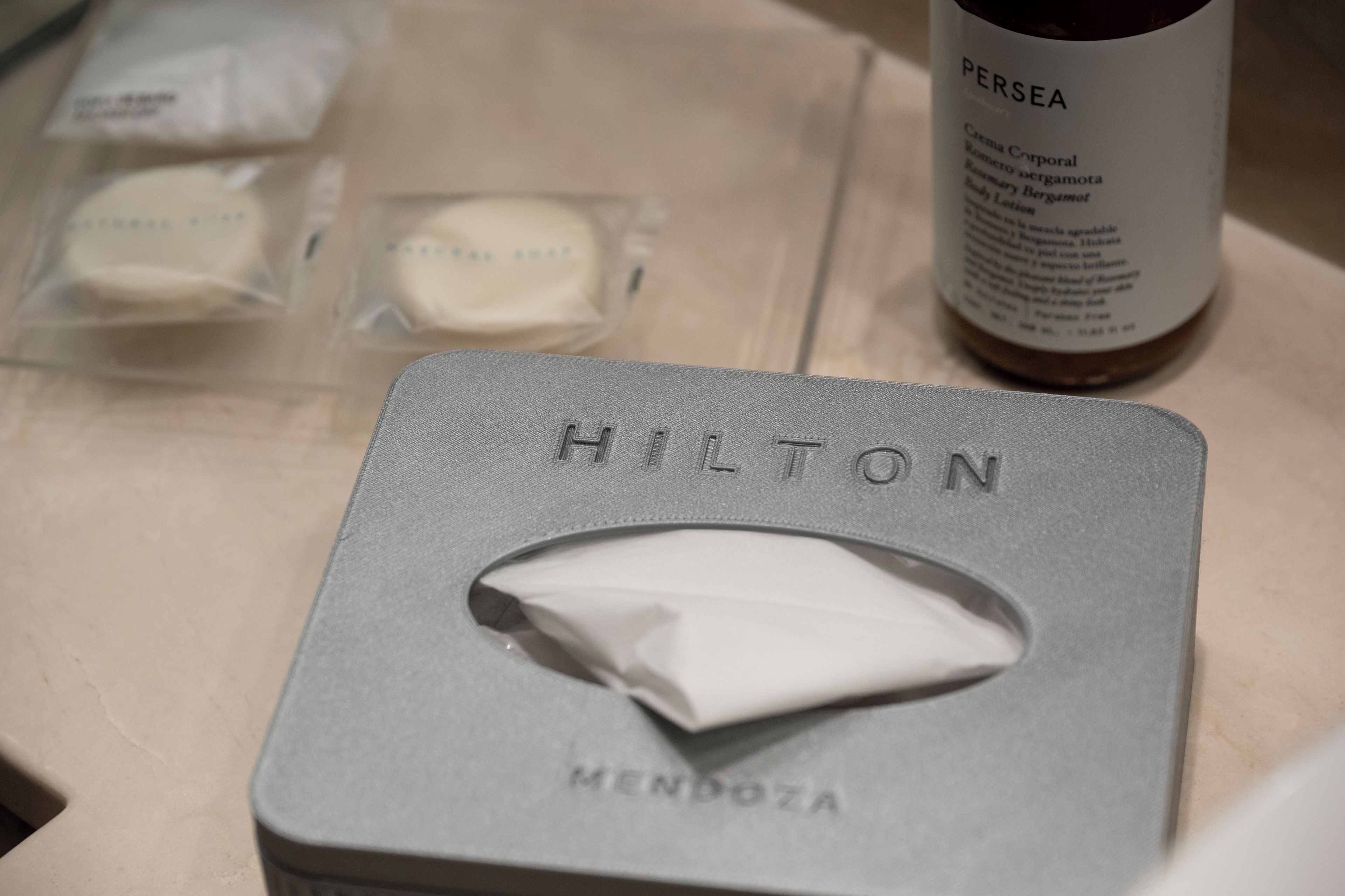 Une chambre d’hôtel dotée d’un minibar bien organisé, comprenant une bouteille de lotion corporelle Persea et une boîte de produits de toilette de la marque Hilton.