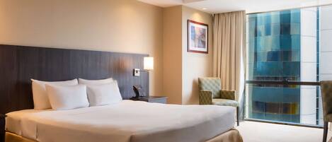 A hotel room with a large bed, a bedside table with a lamp, a chair, and a view of a building through a glass door.
