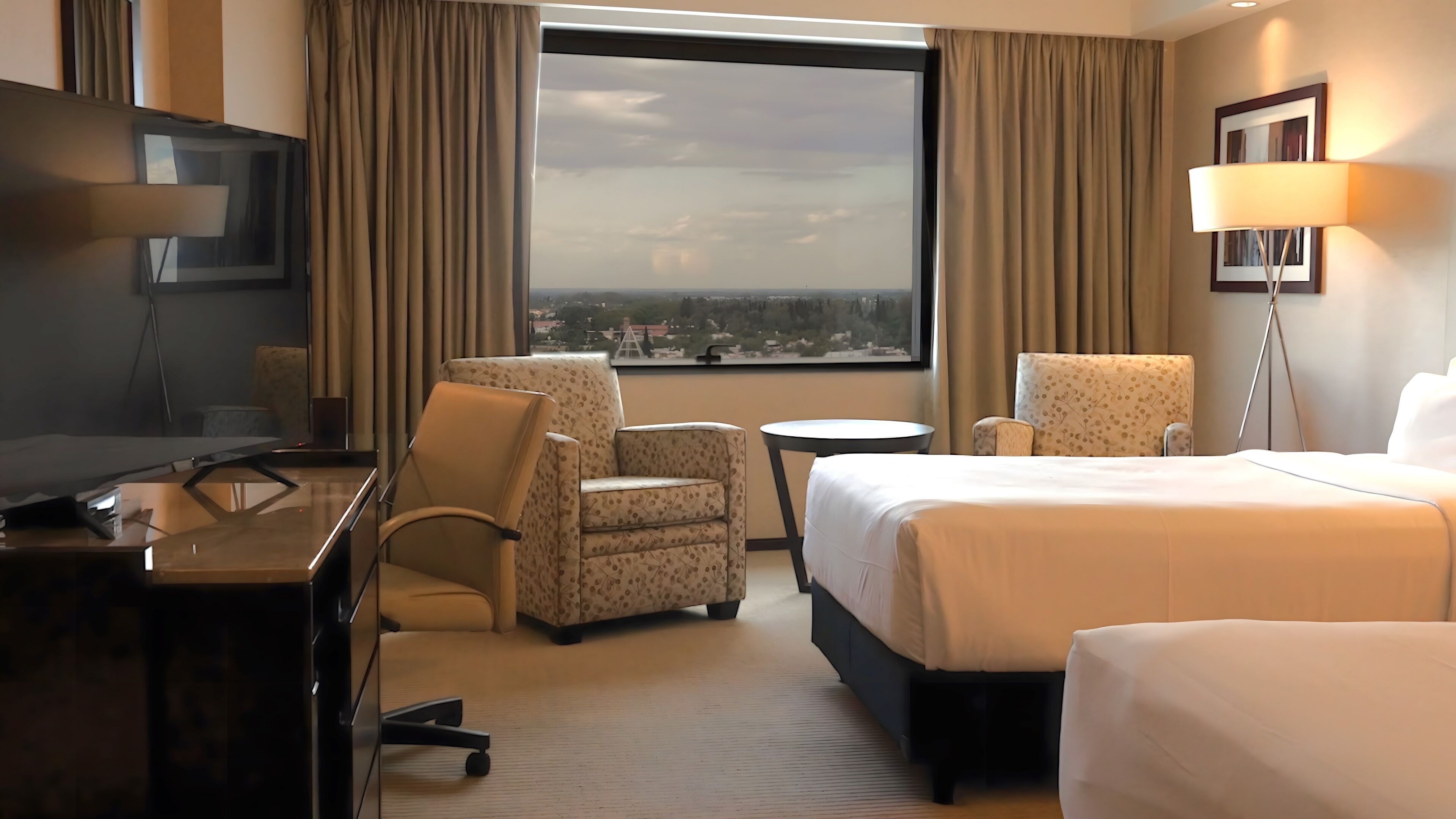 A hotel room with a bed, two armchairs, a desk with a TV, a small table, and a large window offering a city view.
