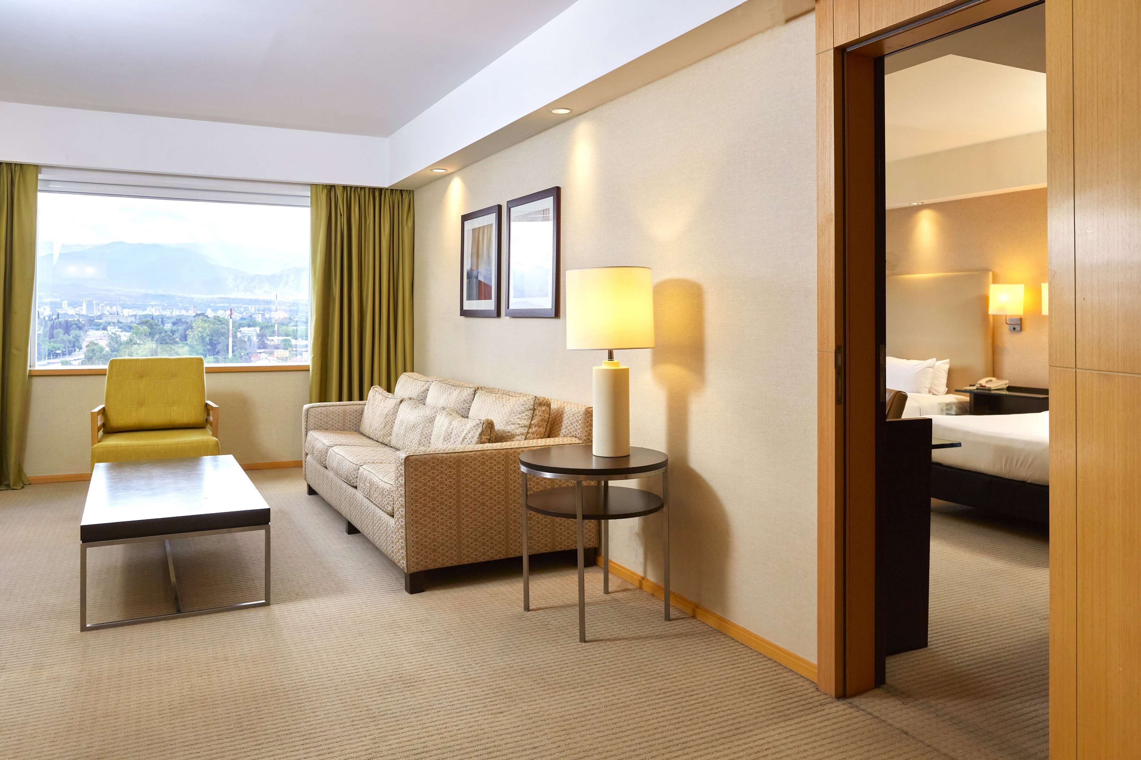 A hotel room with a sofa, a yellow armchair, a small table, a lamp, and a bed.