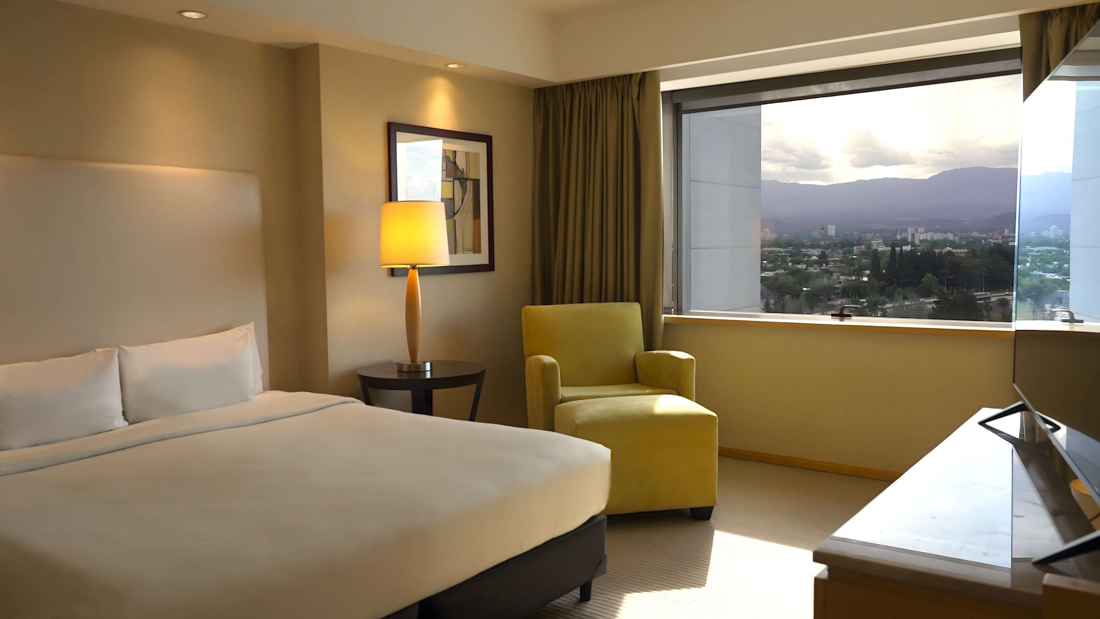 Ein Hotelzimmer mit einem Bett, einem Sessel, einem Schreibtisch, einer Lampe, einem gerahmten Bild und einem Fenster mit Blick auf die Stadt.