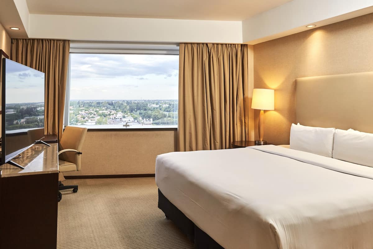 A hotel room with a bed, a desk with a flat-screen TV, a chair, a lamp, and a view of a cityscape through large windows.