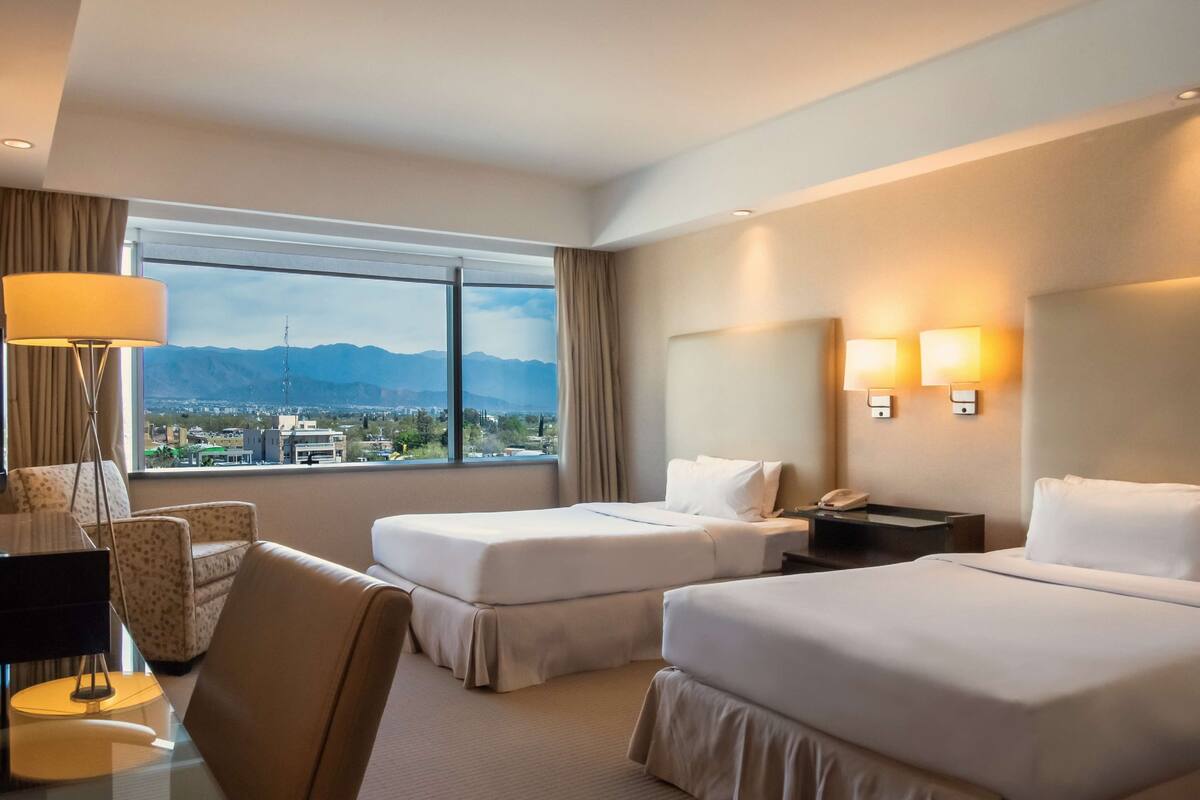 A hotel room with two beds, a desk, a chair, a lamp, and a view of mountains and a city.
