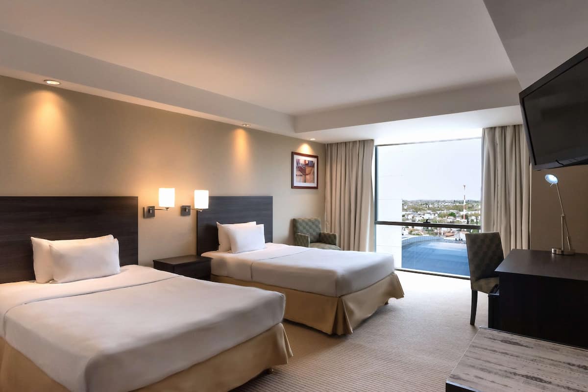 A hotel room with two beds, a desk, a TV, and a view of the city.