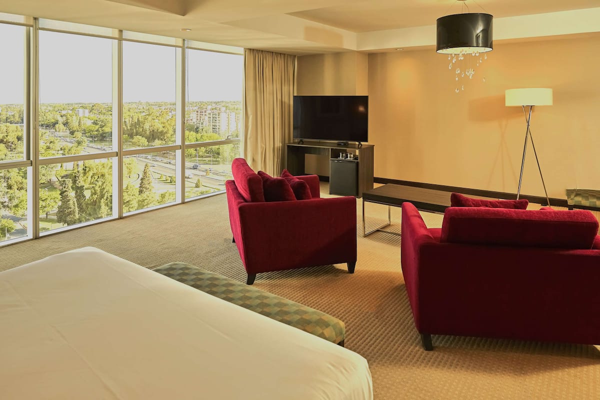 A hotel room with a large bed, two red armchairs, a TV, and a view of a cityscape through large windows.