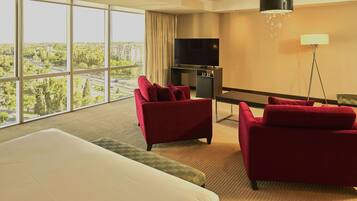 Habitación de hotel con una cama grande, dos sillones rojos, un televisor y vistas a un paisaje urbano a través de amplias ventanas.