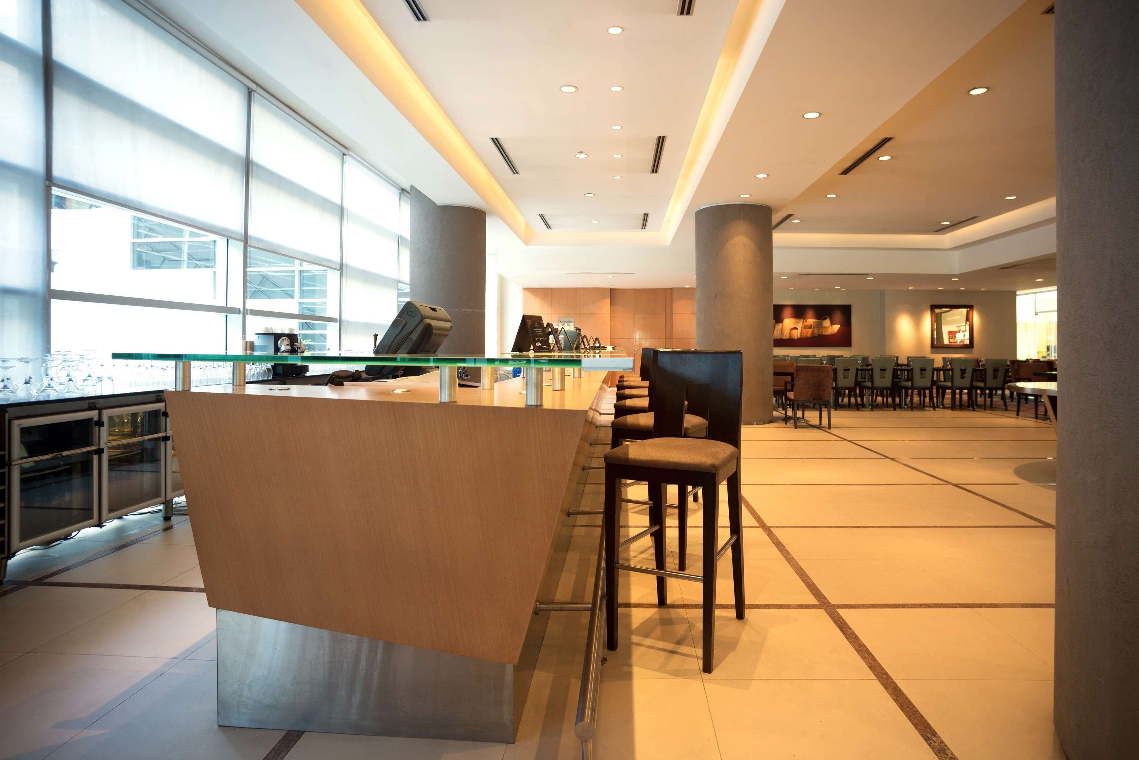 A modern hotel lobby with a glass reception desk, seating area, and large windows providing natural light.