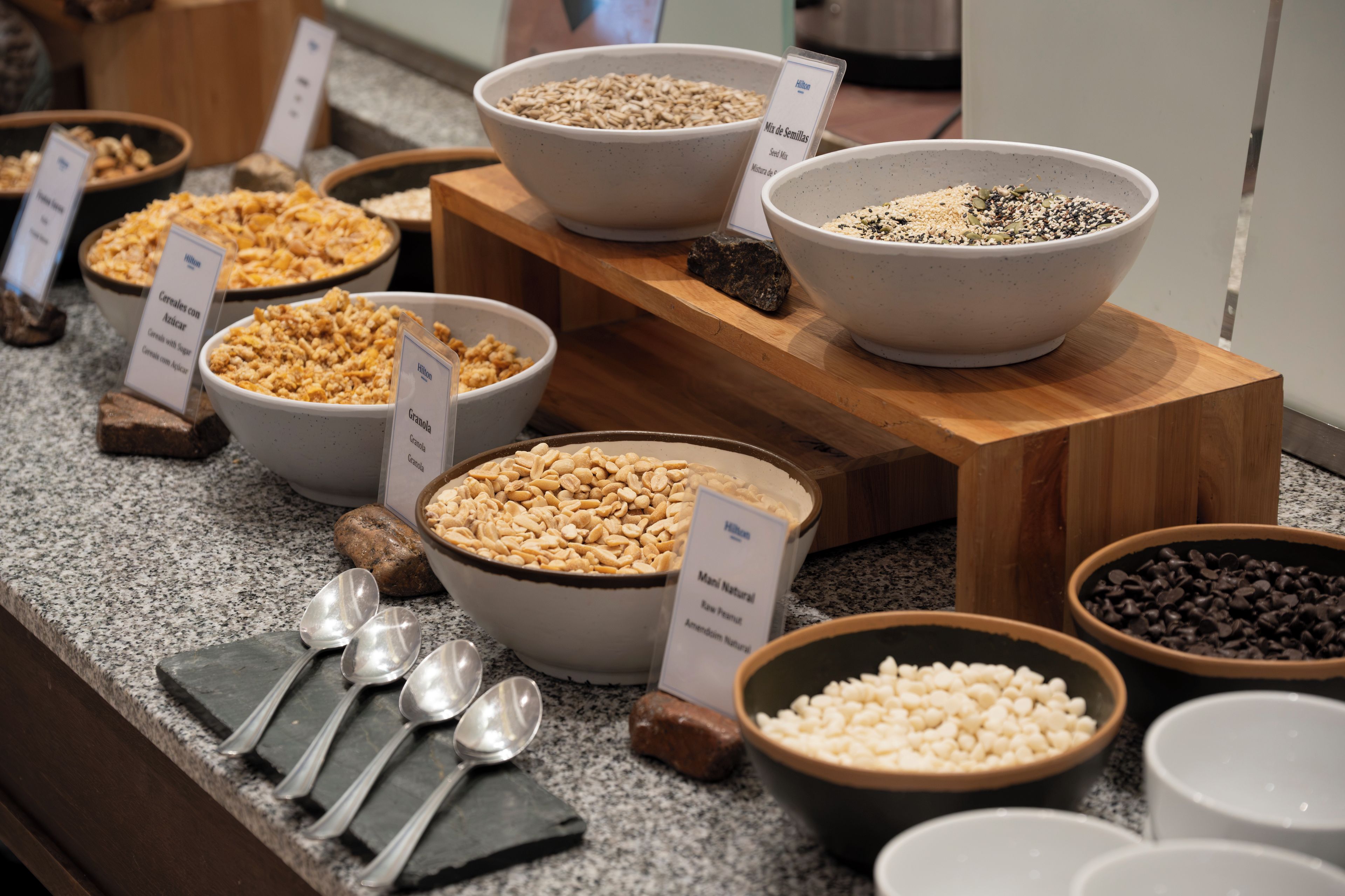 Un bufé con diversas opciones de comida, incluyendo cereales, granola y nueces, con utensilios de servicio y etiquetas que indican el tipo de alimento.