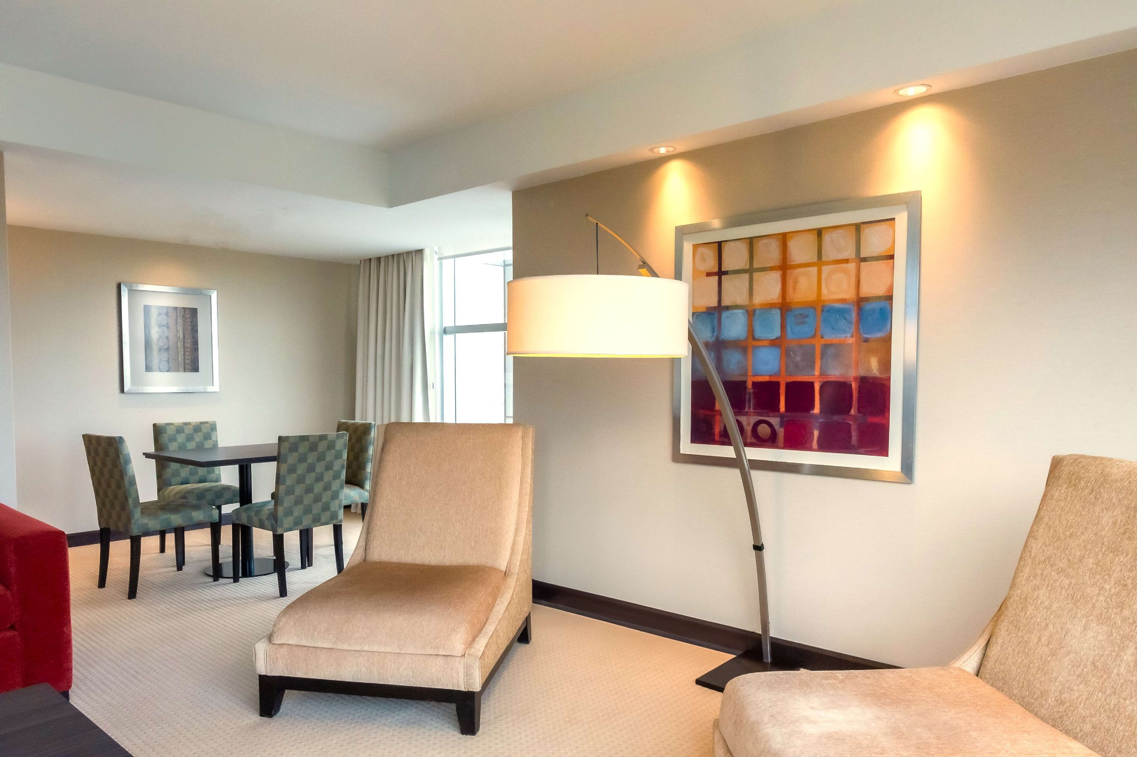 A hotel room with a beige armchair, a dining table with checkered chairs, a modern floor lamp, and a colorful abstract painting on the wall.