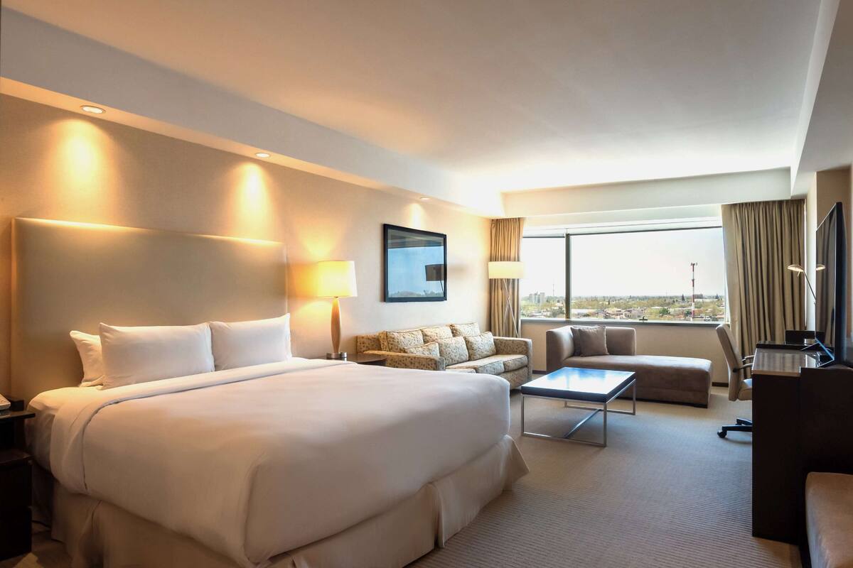 A hotel room with a large bed, a sofa, a desk, and a view of the city.