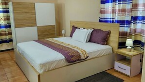 A neatly made bed with a patterned blanket, a bedside table with a lamp, and a wardrobe with a wooden panel design. - Serene Hostel Accra Bunk Bed Mixed dormitory (Adenta Municipality)