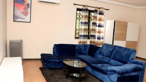 A living room with a blue sofa, a small black coffee table, a white TV stand, and a large window with striped curtains. - Serene Hostel Accra Bunk Bed Mixed dormitory (Adenta Municipality)