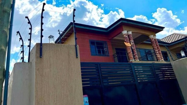 A two-story house with a red and beige facade, a black gate, and a clear blue sky with clouds. - Serene Hostel Accra Bunk Bed Mixed dormitory (Adenta Municipality)