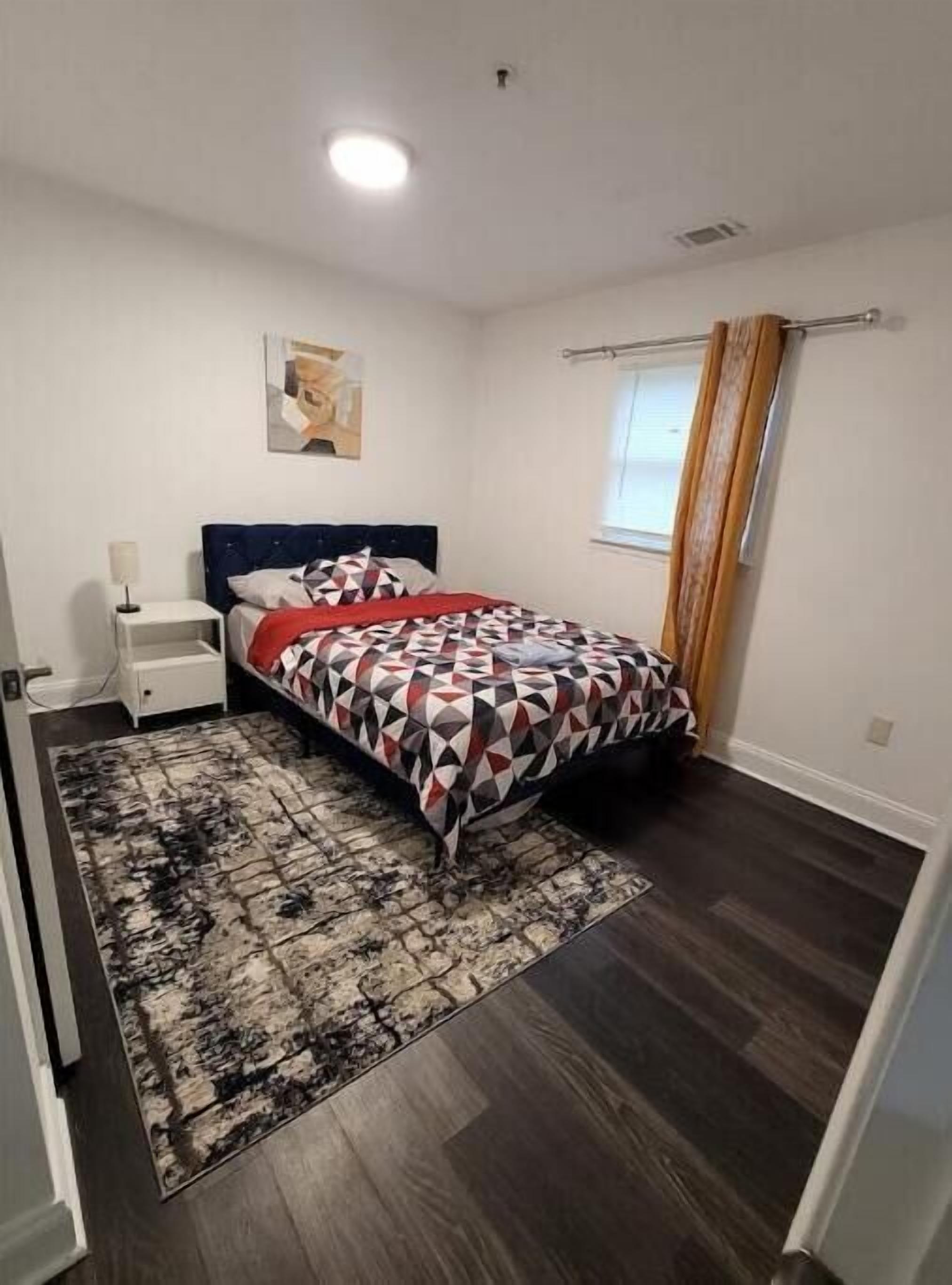 Un dormitorio con cama, mesita de noche, lámpara, un cuadro en la pared y ventana con cortinas.