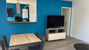 A modern living room with a blue accent wall, a wooden dining table with four chairs, a TV on a wooden stand, and a white door. - Spacious T3 with balcony - close to town center (Touques)