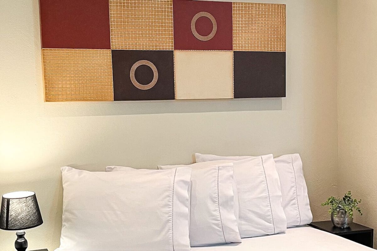 A neatly made bed with white linens and a brown throw blanket, a bedside table with a lamp, and a wall-mounted artwork above the bed.