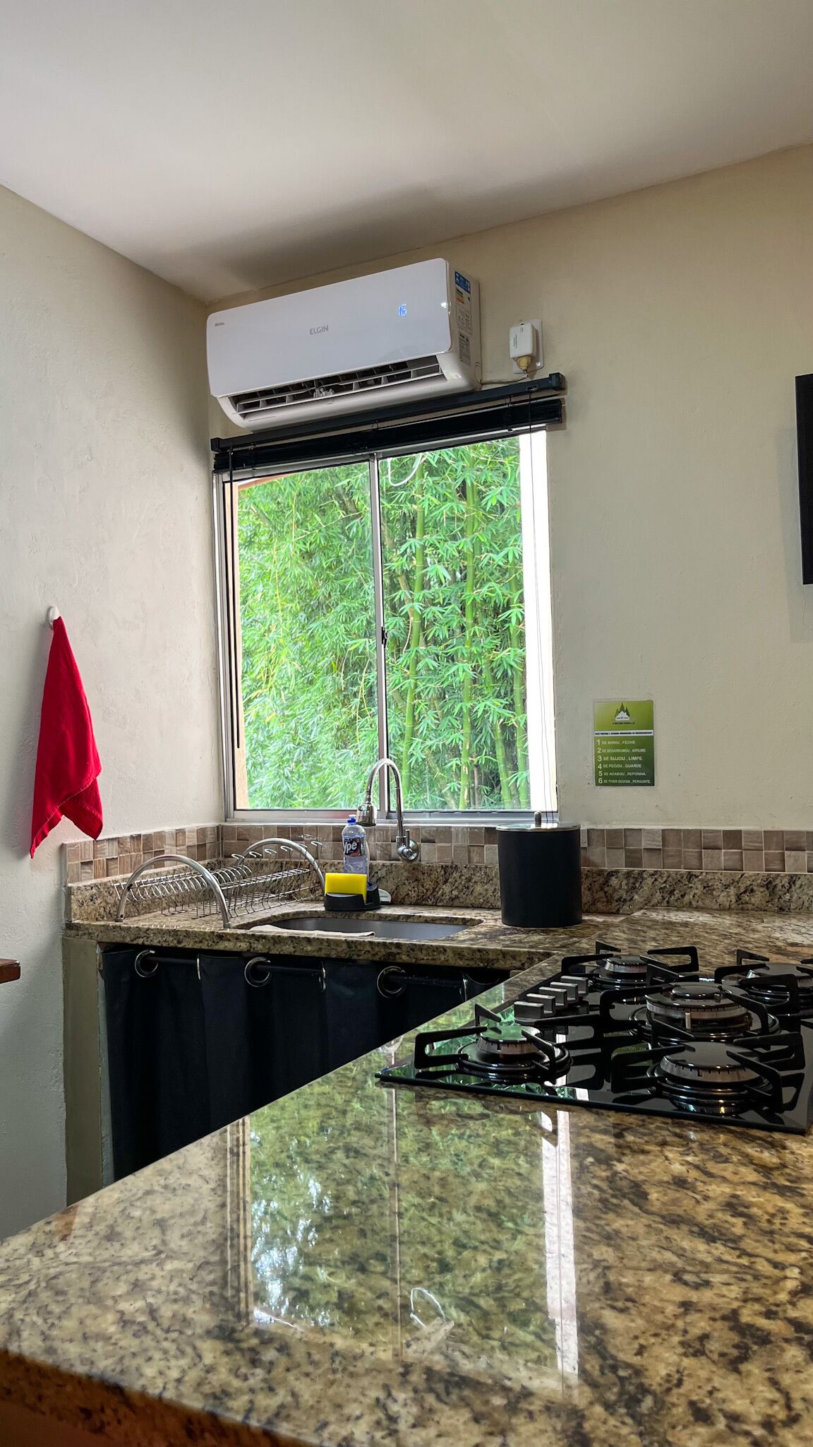 Une cuisine équipée d’une cuisinière à gaz, une fenêtre donnant sur un paysage verdoyant, une serviette rouge accrochée au mur et un appareil de climatisation placé au-dessus de la fenêtre.