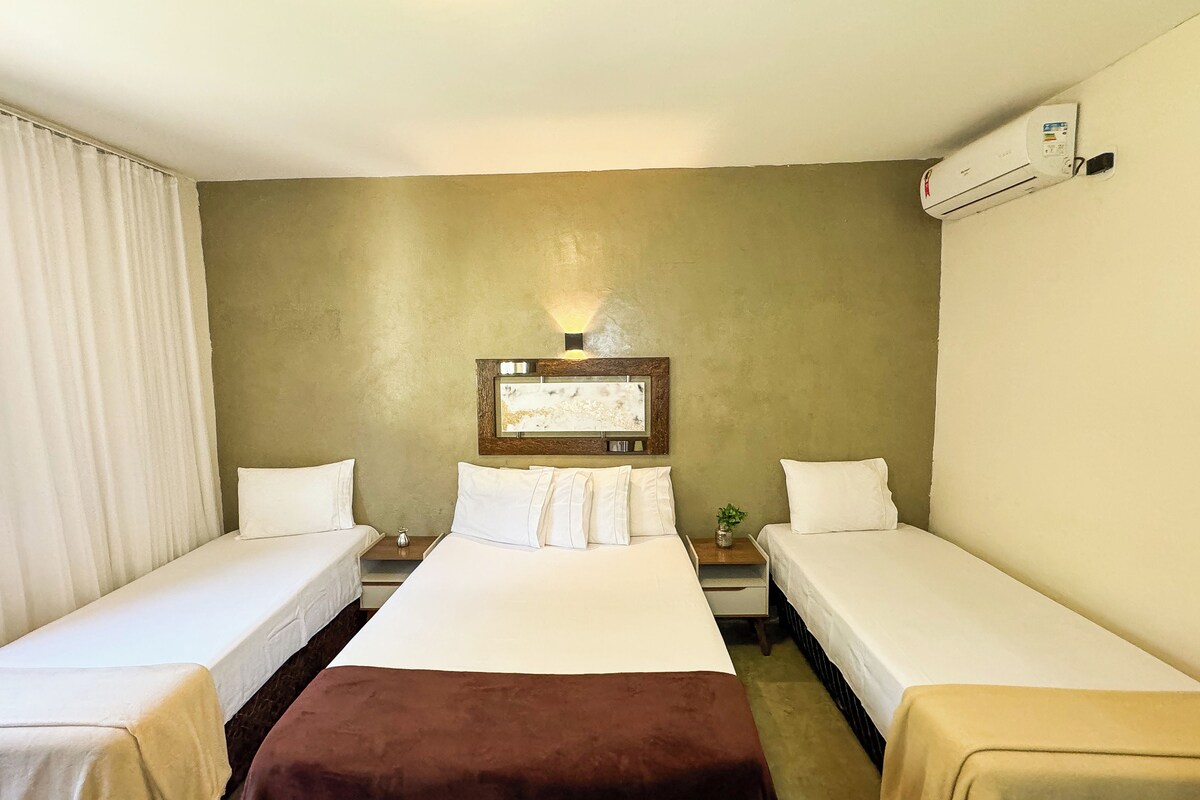 A hotel room with two single beds, a large bed, a nightstand with a lamp, and an air conditioning unit.