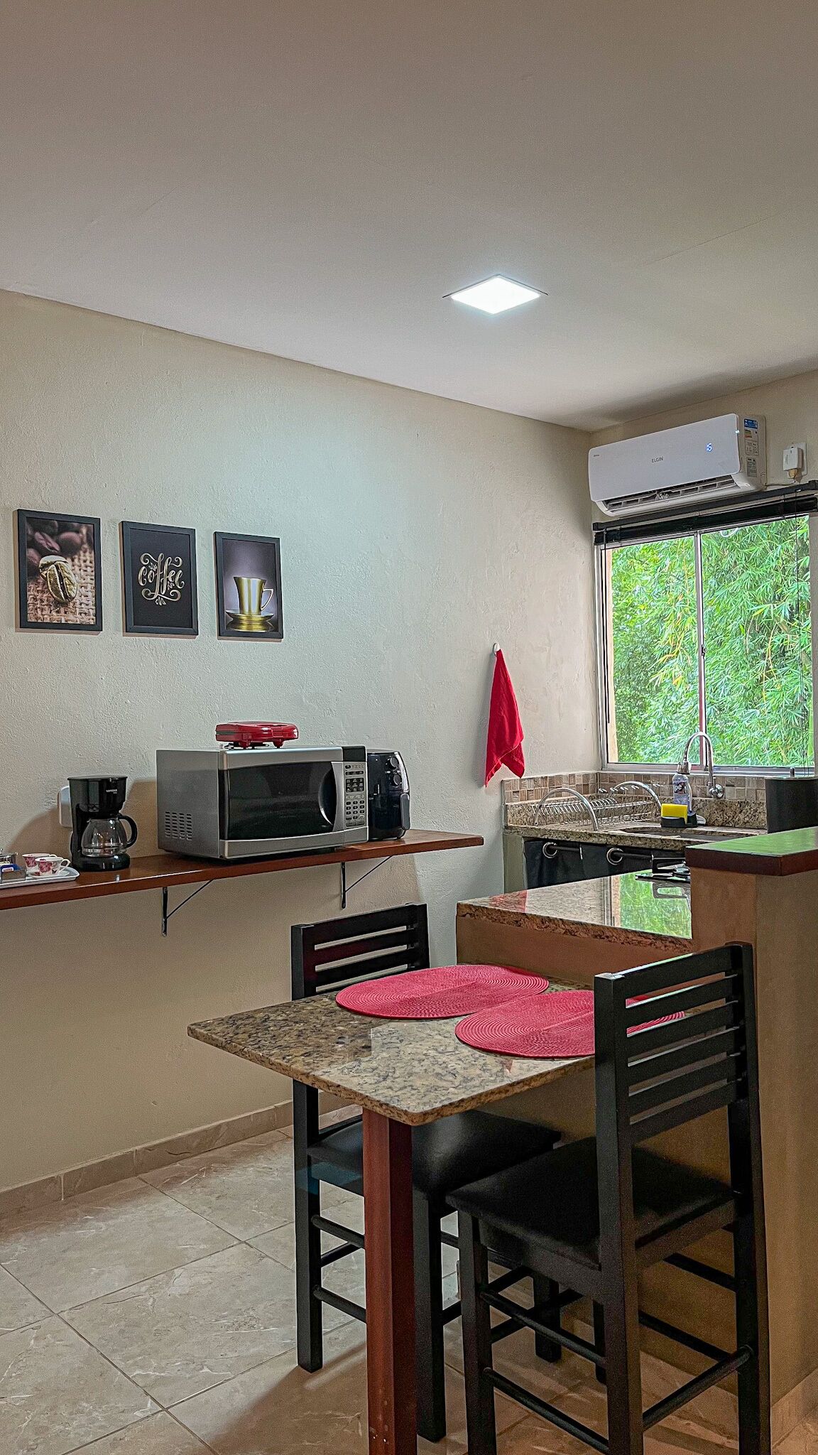 A compact kitchen with a microwave, coffee maker, and a small dining table with chairs. There are framed pictures on the wall and a window with a view of greenery.