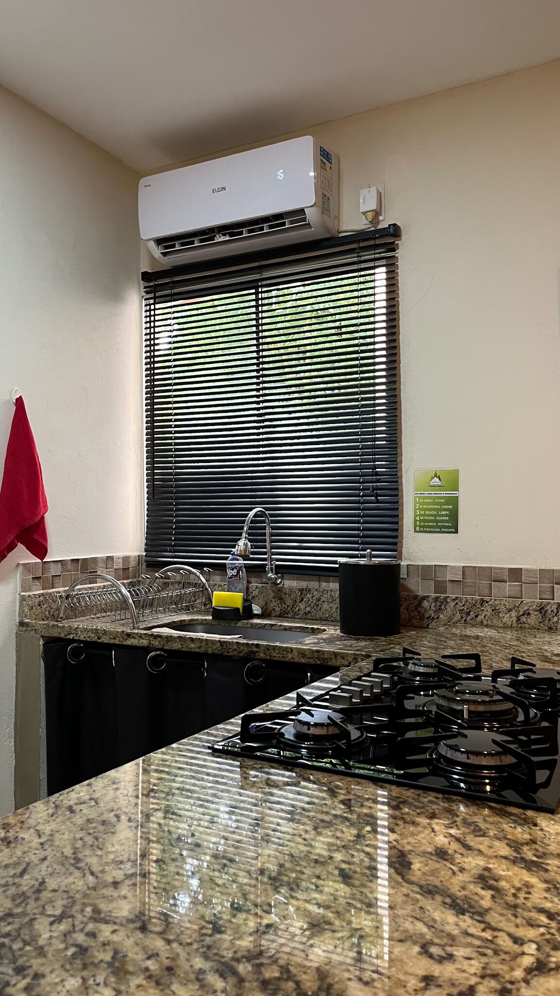 Een keuken met een gasfornuis, een spoelbak, een raam met jaloezieën en een airconditioning.