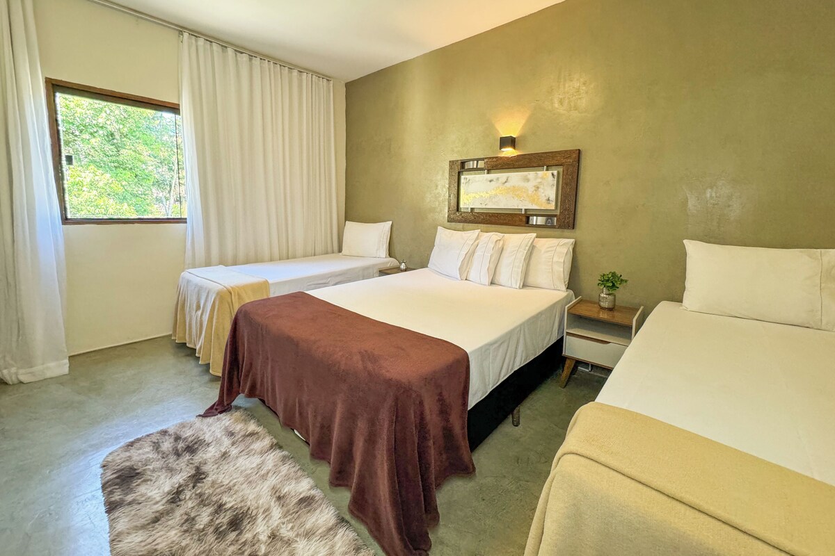 Une chambre d’hôtel avec deux lits simples, un grand lit, une petite table avec une plante et une fenêtre avec des rideaux.