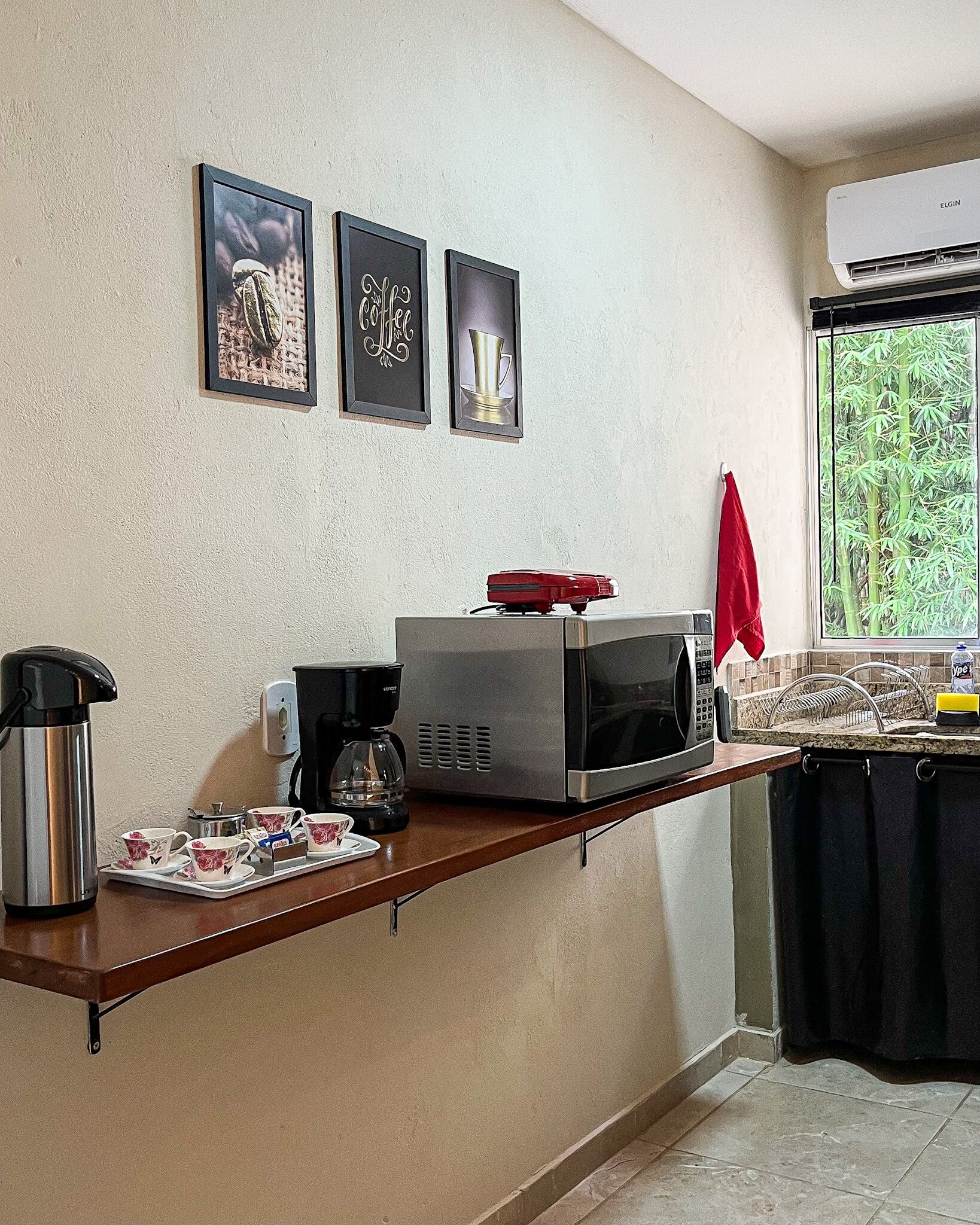 A kitchenette with a microwave, coffee maker, and tea kettle on the counter. There are framed pictures on the wall and a window with a view of bamboo.