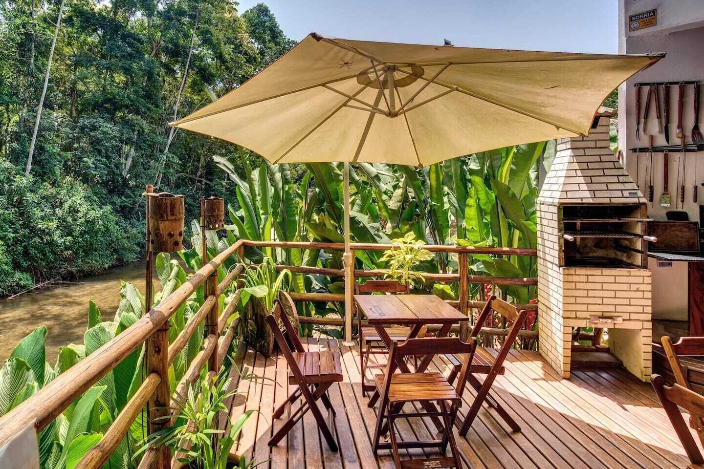 Una terraza de madera con una mesa y sillas bajo un gran paraguas, con vista a un río y follaje denso.