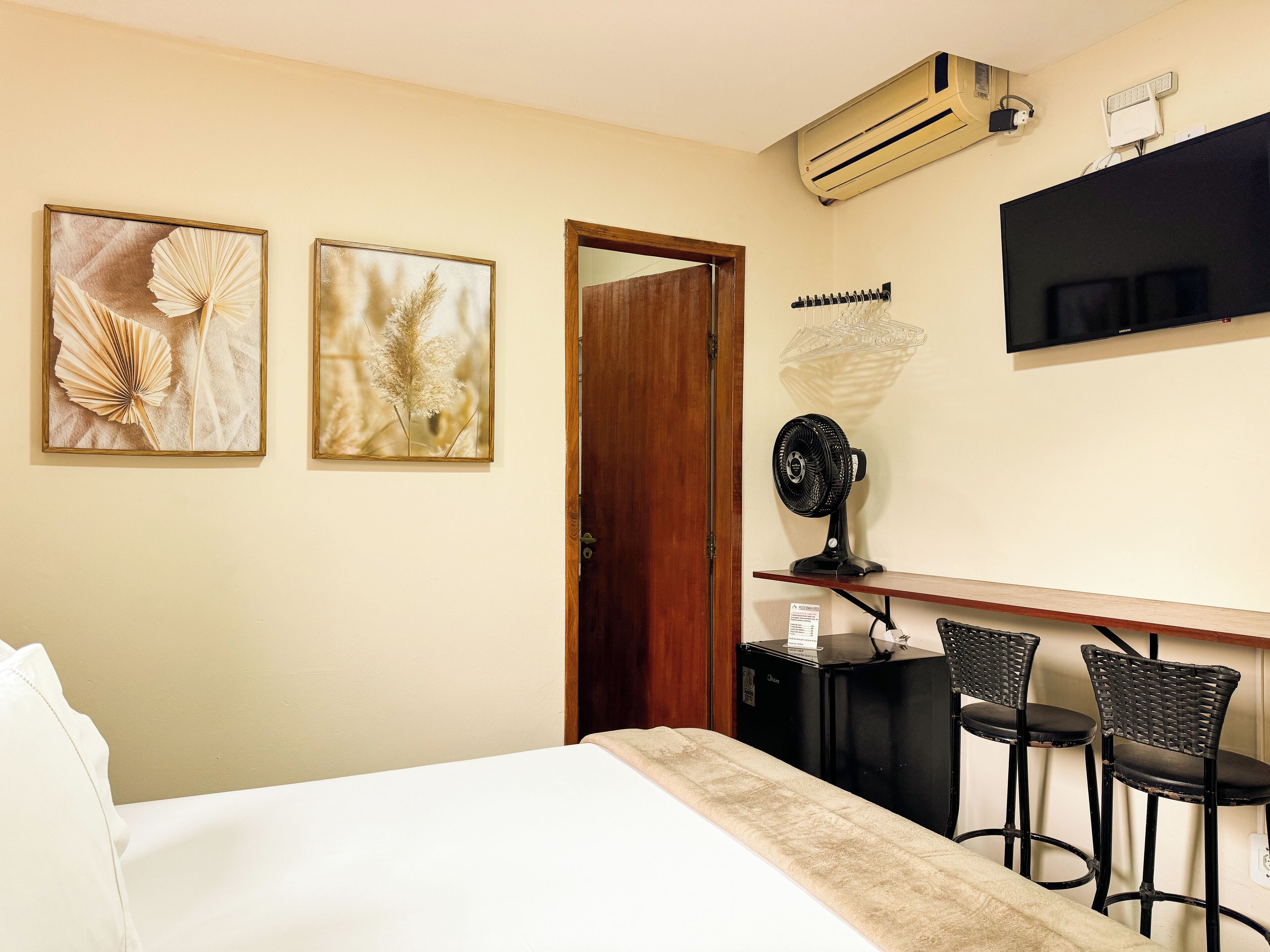 A hotel room with a bed, two wall-mounted artworks, a desk with a chair, a fan, and a TV.