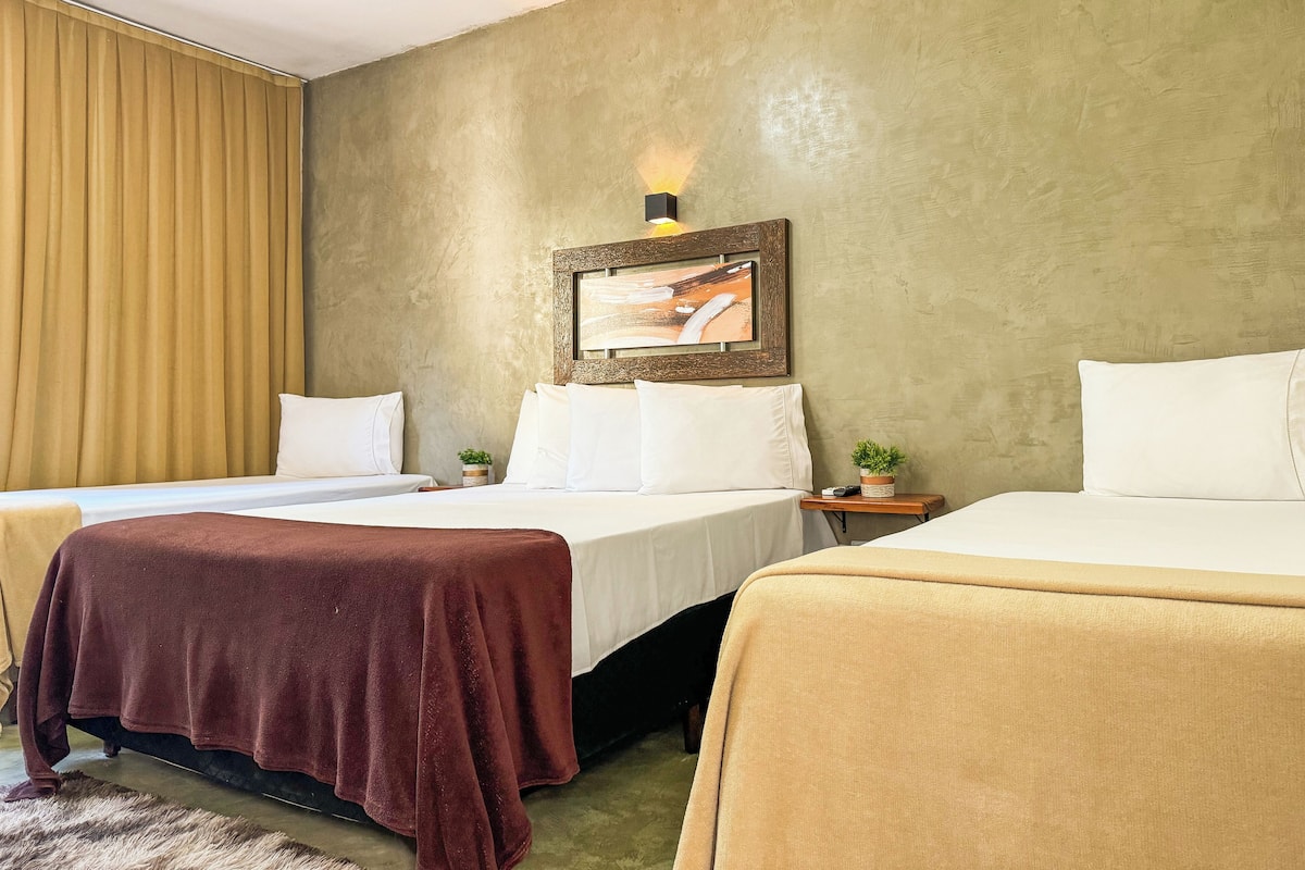A hotel room with two beds, a large window with curtains, a painting on the wall, and a small table with a plant.