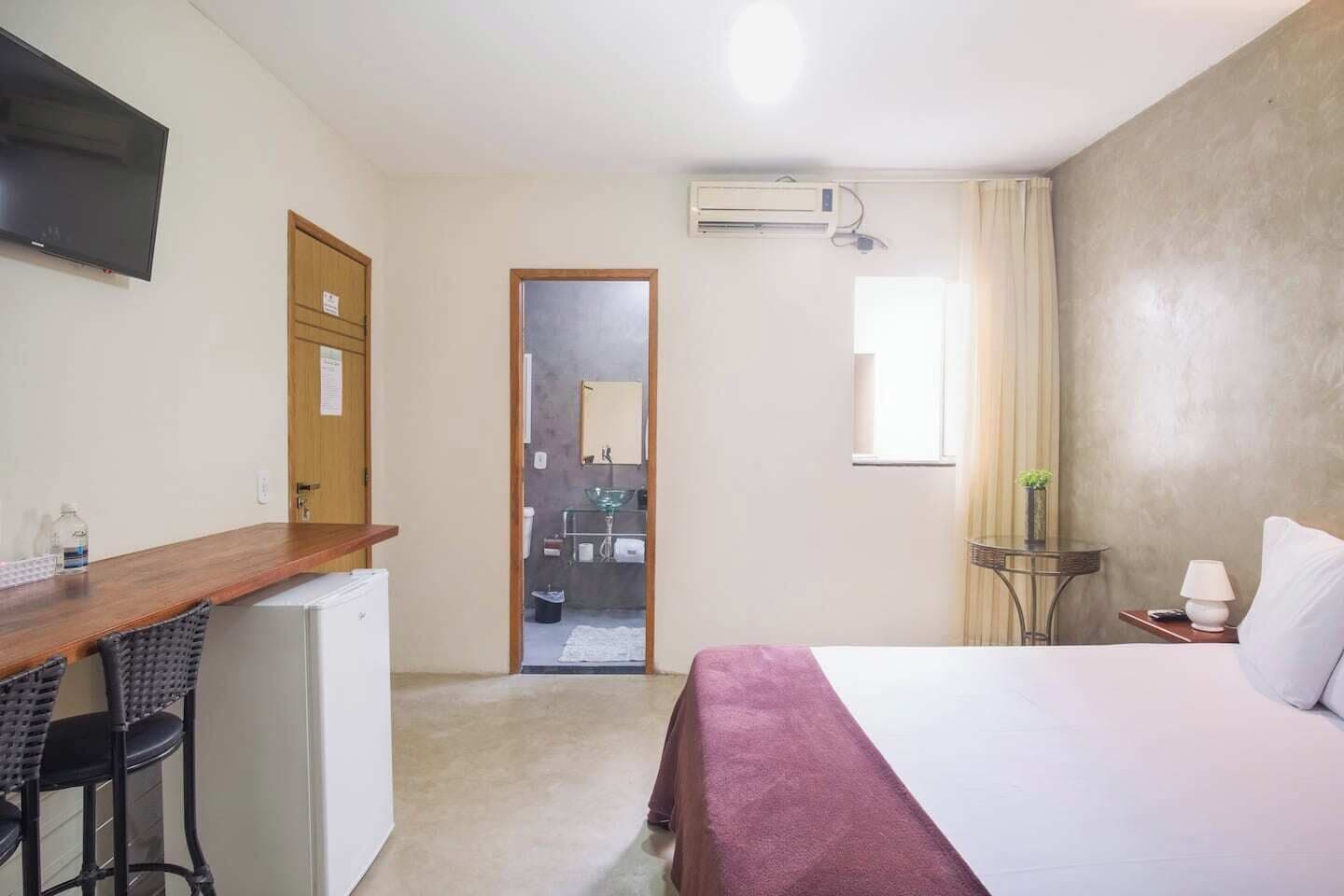 Et hotelværelse med seng, skrivebord, stol, et lille bord med lampe og et klimaanlæg.