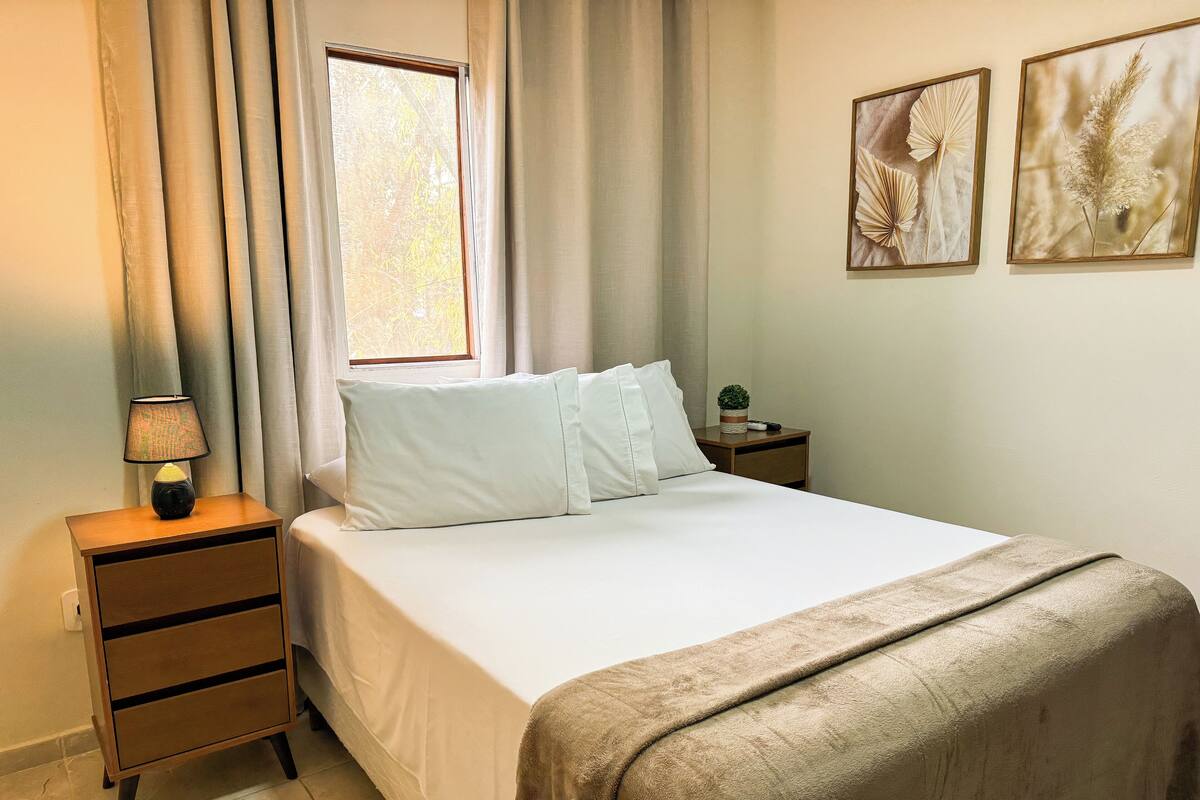 A neatly made bed with white linens, a bedside lamp, and a small bedside table with a plant and a remote control.