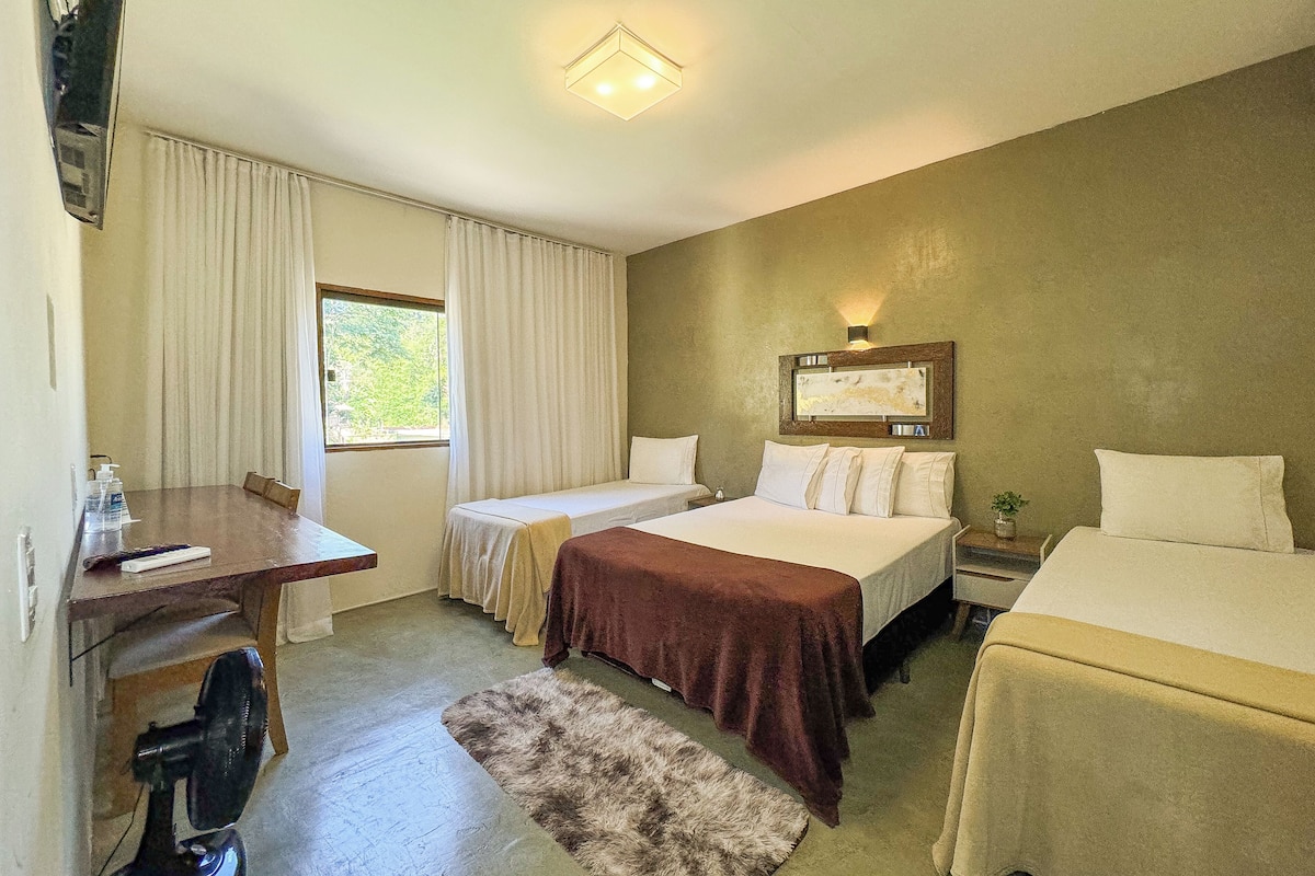 A hotel room with two beds, a desk, a chair, a small table, a fan, and a window with curtains.
