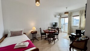 Et hotelværelse med en seng, et spisebord med stole, et fjernsyn og en balkon med gardiner.