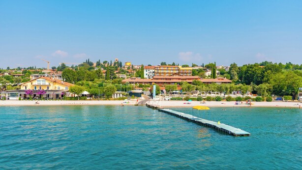A coastal town with a pier extending into the water, surrounded by buildings and greenery. - Mobilehome with terrace, pool and air conditioning (Padenghe sul Garda)