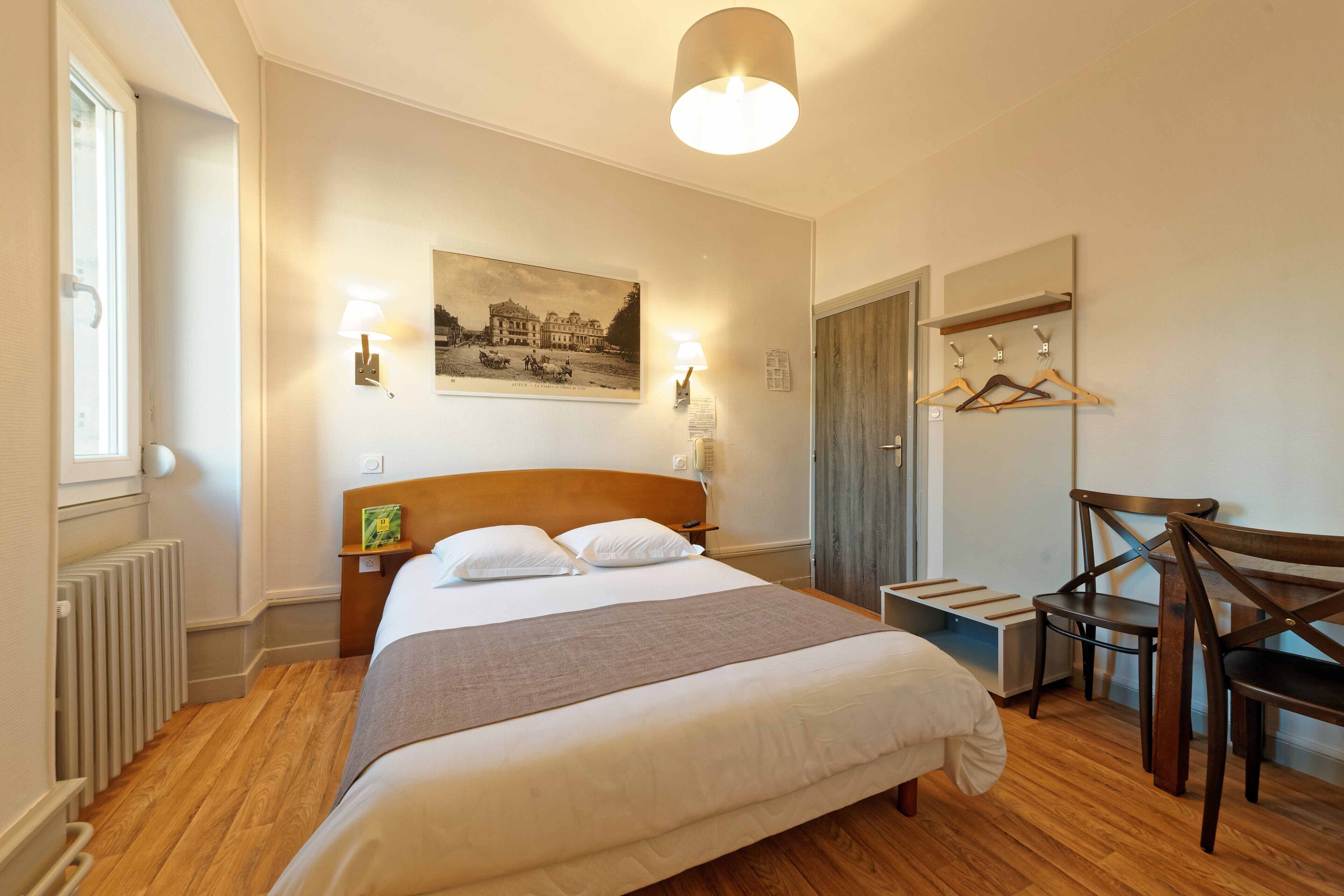 Une chambre bien rangée, avec un grand lit, des lampes de chevet, une tête de lit en bois, un radiateur, une chaise en bois, un porte-manteau et un tableau encadré au mur.