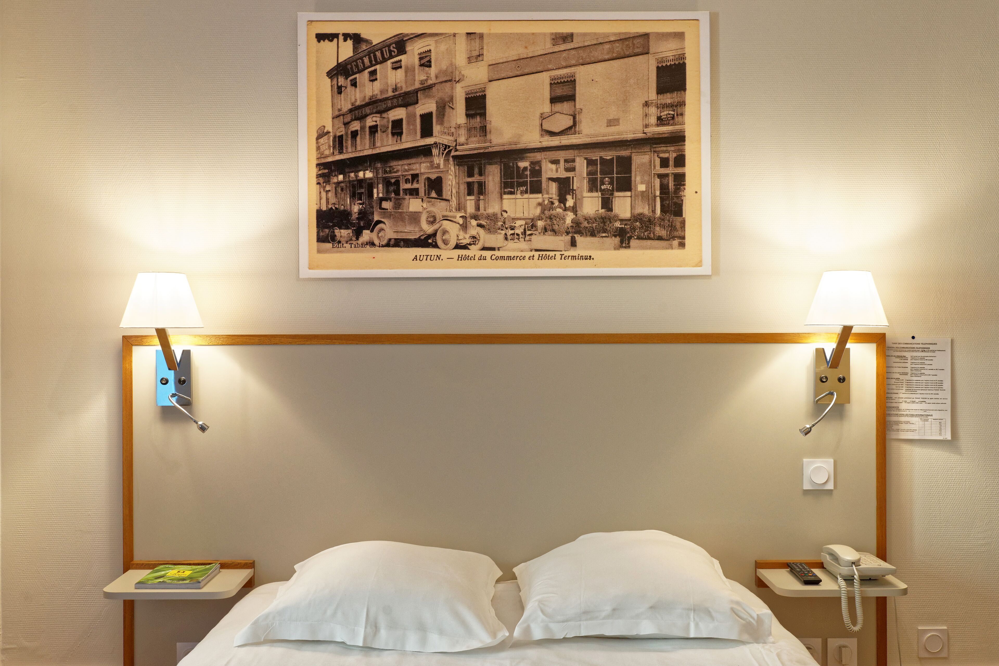 Ein Hotelzimmer mit einem Bett, zwei Nachttischlampen, einem Nachttisch mit einem Buch und einem Telefon, einem gerahmten Foto an der Wand und einem Holzkopfende.